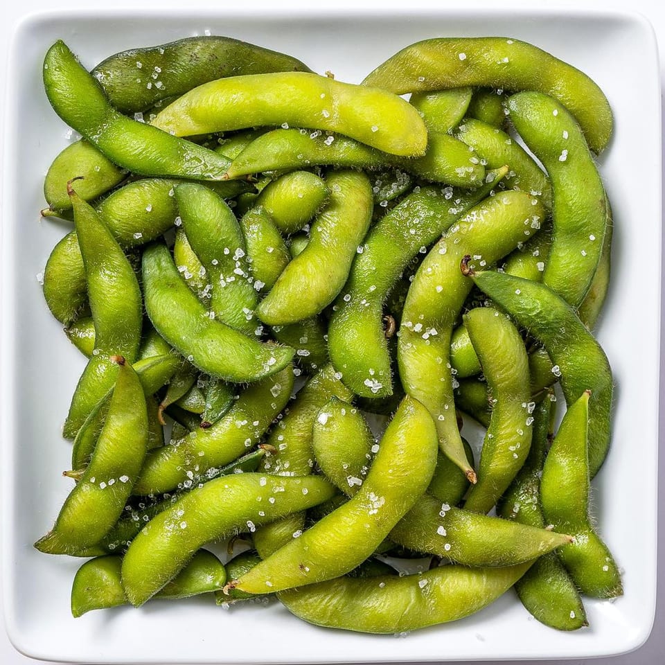 A white square bowl filled with vibrant green edamame pods, lightly sprinkled with coarse sea salt.