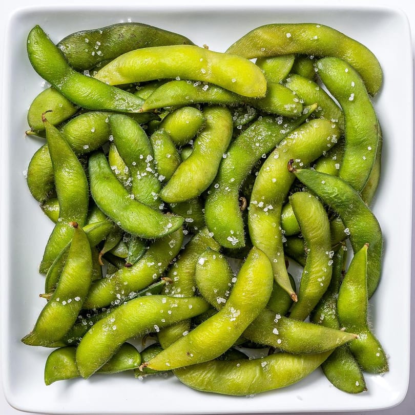 A white square bowl filled with vibrant green edamame pods, lightly sprinkled with coarse sea salt.