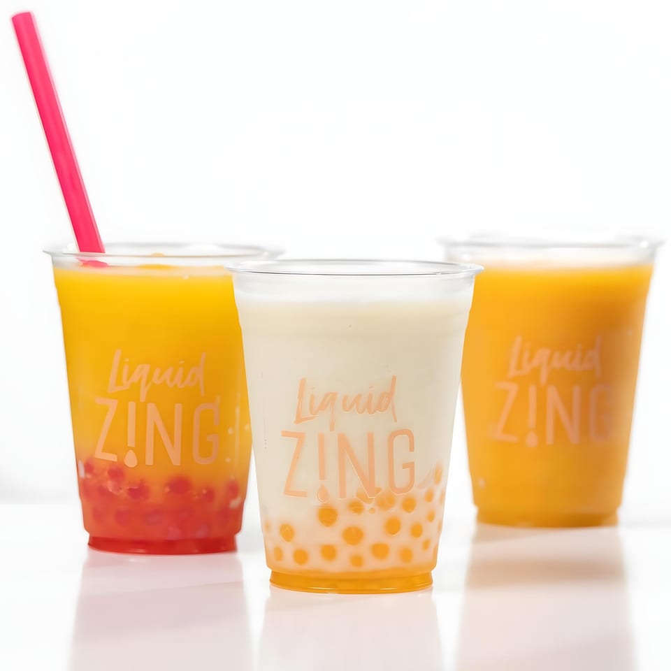 Three Zing bubble tea drinks, two orange and one white, with colorful popping boba on a white surface.