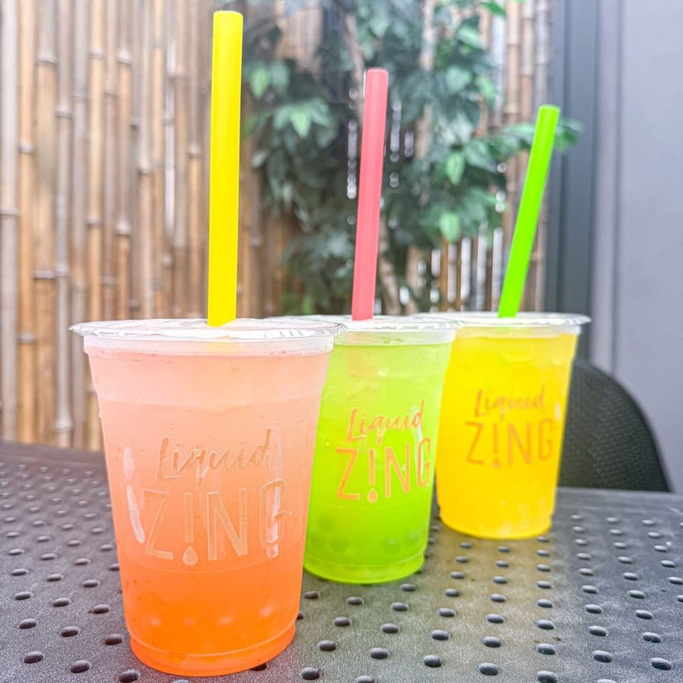 Three colorful Zing drinks with straws, orange, green, and yellow, on a dark textured table.